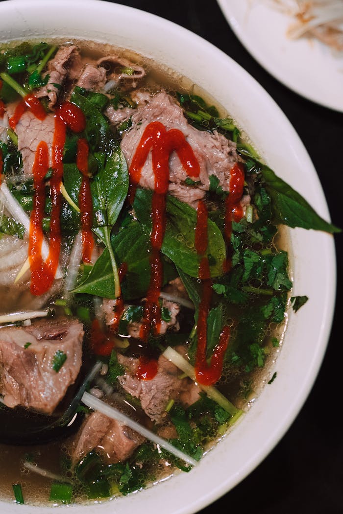 A close-up shot of Vietnamese pho with fresh herbs, beef, and hot sauce.